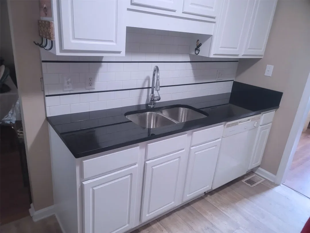 Kitchen counter with white cabinets, black countertop, stainless steel sink, white Subway tile backsplash, and a dishwasher.