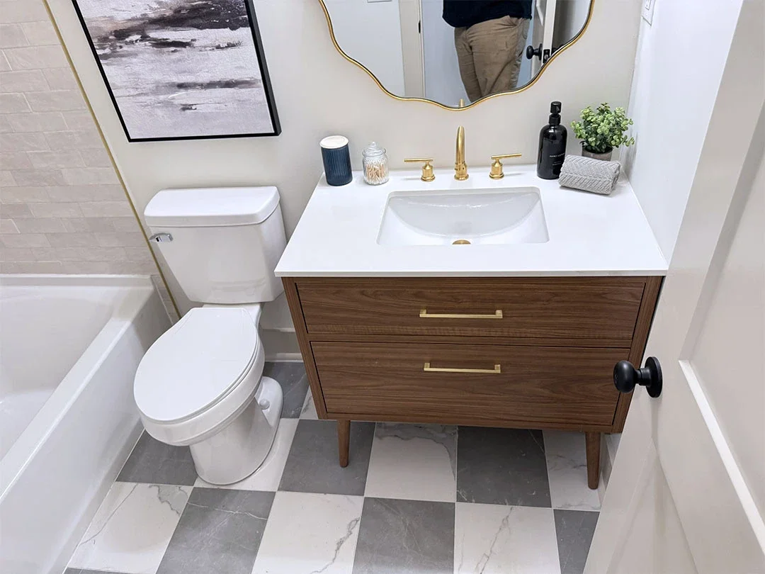 A modern bathroom with a white toilet, a wooden vanity with gold handles, a white countertop, a rectangular sink, gold faucet, a black soap dispenser, a small potted plant, and decorative items. A mirror hangs above the vanity, and a framed abstract artwork is on the wall.