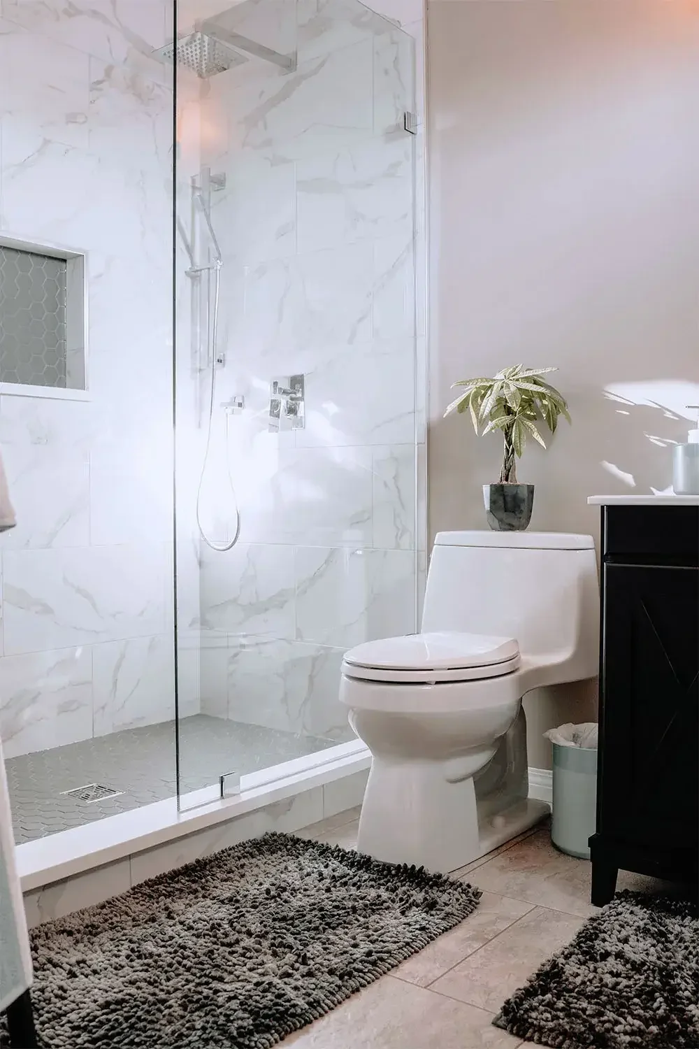 A modern bathroom with a glass-enclosed walk-in shower, white marble tiles, a white toilet, a black vanity with a potted plant on top, and dark gray textured rugs on the beige tile floor.