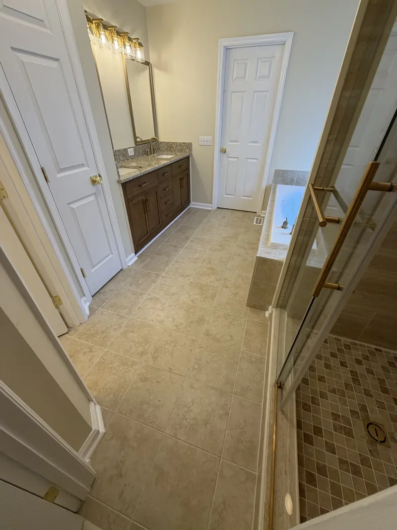 Bathroom with beige tile flooring, granite countertop vanity with a mirror, a closed white door, and a bathtub. Shower area with glass door is partially visible.