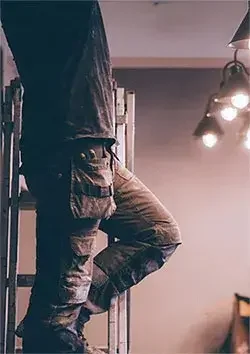 Person in military or tactical clothing standing on a ladder, with a ceiling light fixture visible in a room.
