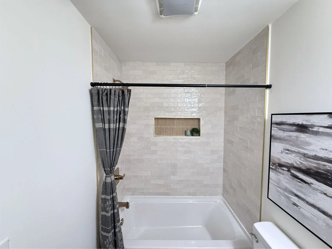 Bathroom with a shower, grey patterned curtain, white tub, white brick tile wall, decorative niche with plant, abstract black and white artwork, air vent on ceiling.