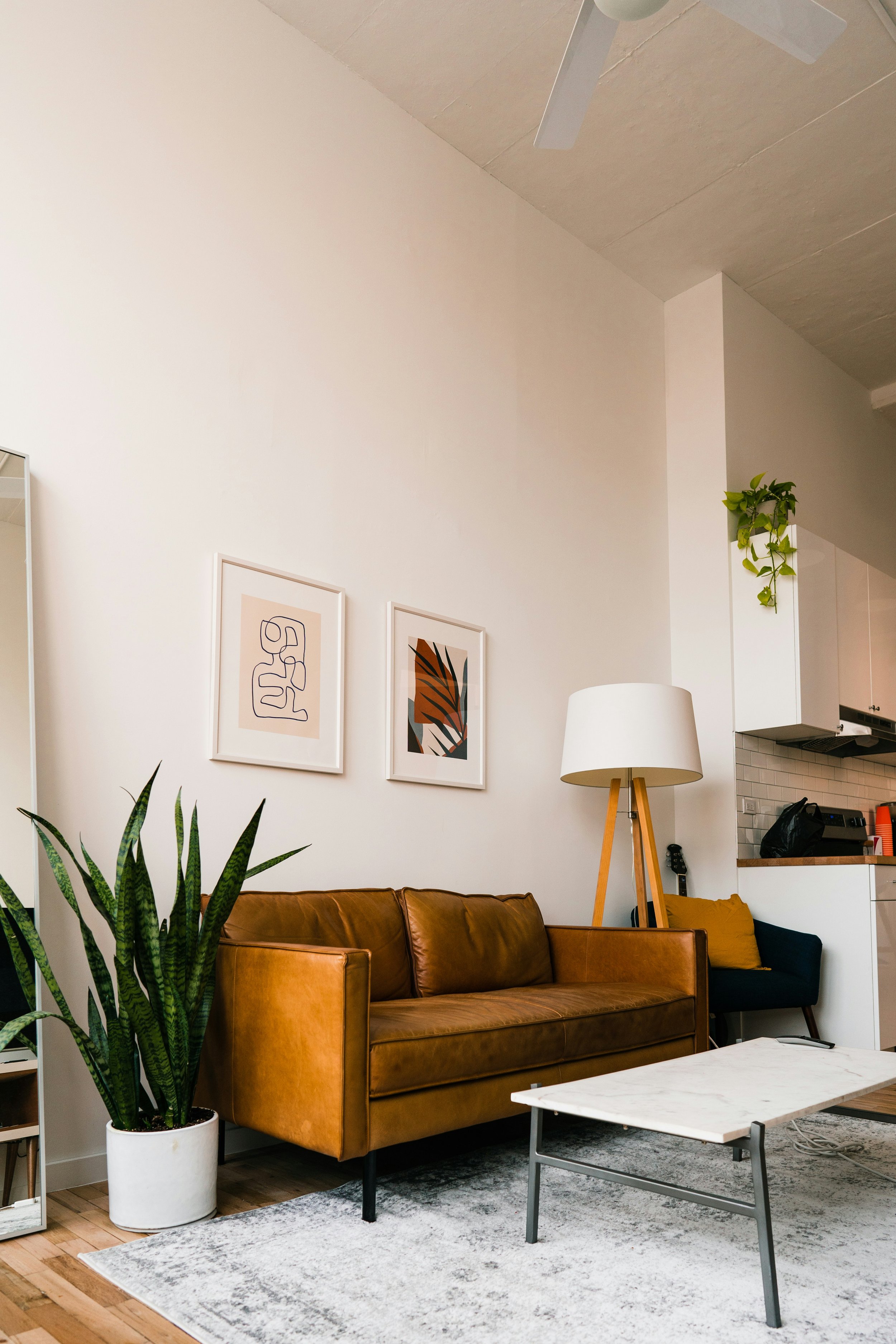 Living room with a tan leather sofa, a white marble coffee table, a tall floor lamp with a wooden tripod base, framed abstract art on the wall, a potted snake plant, and a partial view of a kitchen with white cabinets and black appliances.