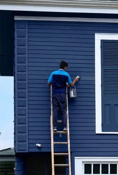 A person painting the exterior wall of a blue house from a ladder.