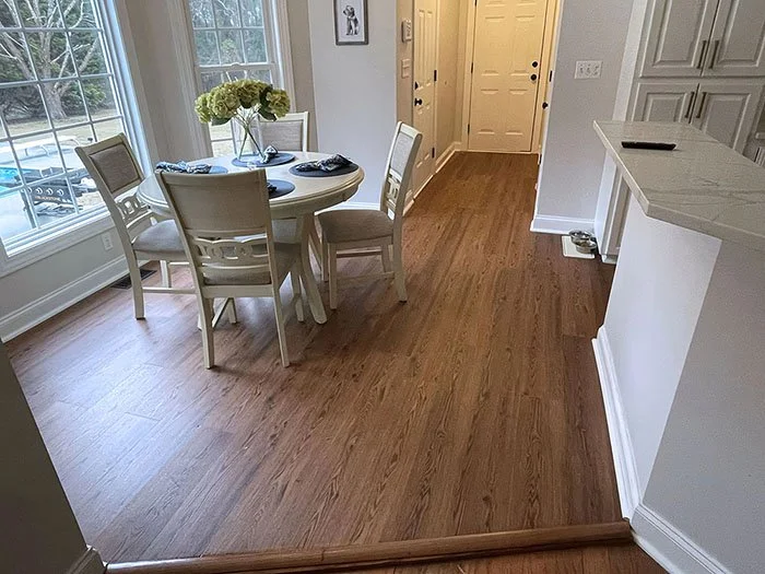 Dining area with a round table, four chairs, a vase of flowers, located near large windows in a bright kitchen with hardwood floors and white walls.