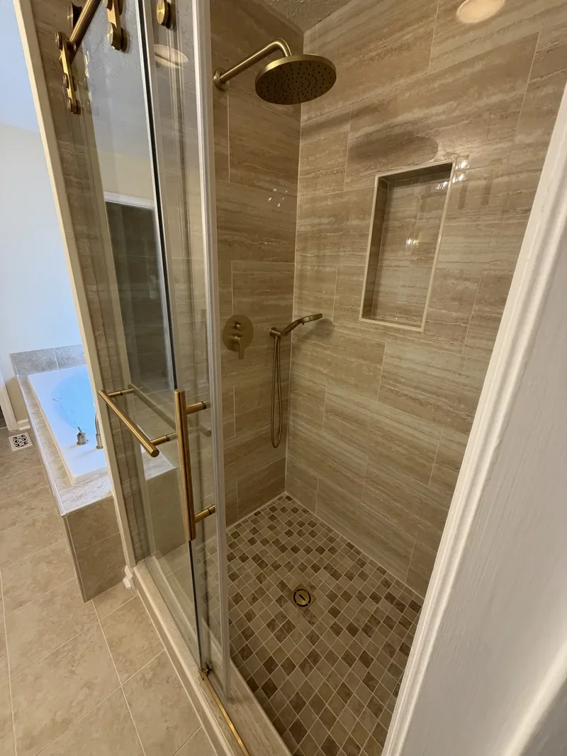 A modern walk-in shower with beige tiled walls and stone floor tiles. It has a rainfall showerhead, a handheld shower, a built-in wall niche, and a glass door with gold handles.