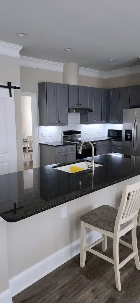 Kitchen with gray cabinets, stainless steel refrigerator, black countertop, white walls, and a white chair at the counter.