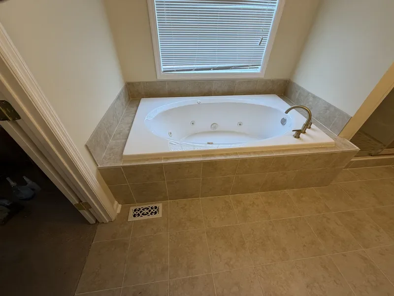 A bathroom corner with a built-in bathtub surrounded by beige tiles, a brass faucet, and a window with closed blinds.
