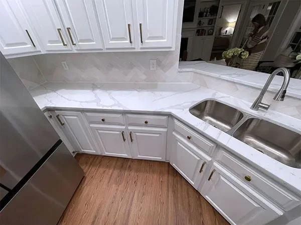 White kitchen cabinets with gold handles, marble countertop, stainless steel double sink, and a stainless steel refrigerator in a modern kitchen.