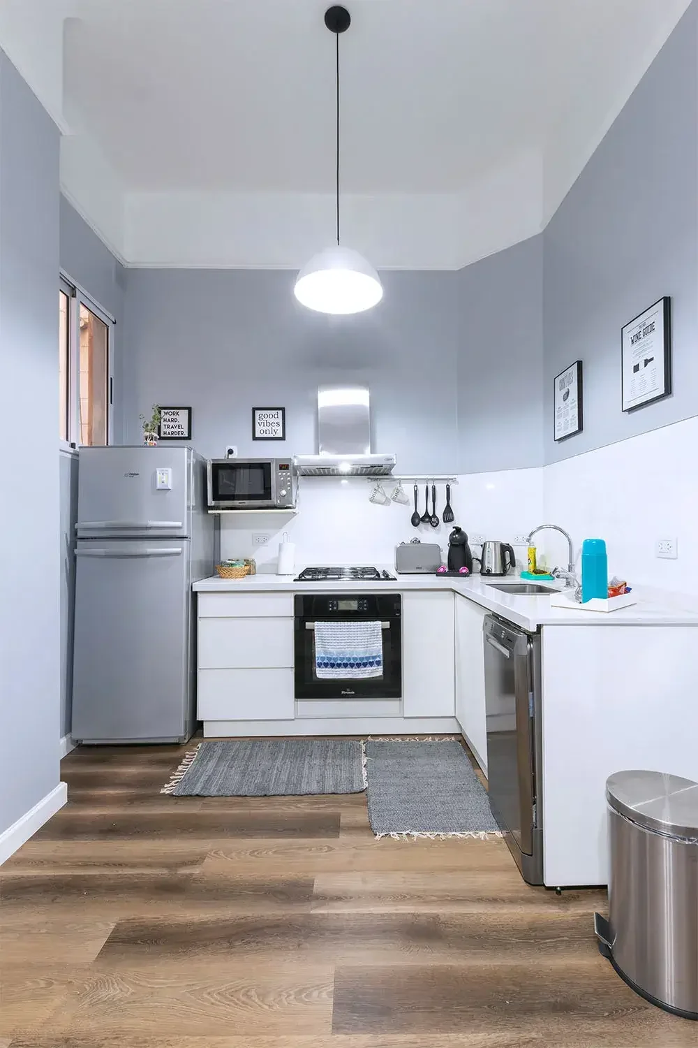 Modern kitchen with white cabinets, gray walls, and stainless steel appliances including a refrigerator, microwave, oven, and dishwasher. Small decor and kitchen essentials are neatly arranged, with hanging utensils and framed quotes on the wall. Lig