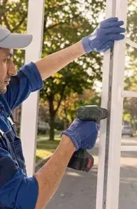 A person wearing a cap and gloves using a power drill to assemble or repair a white wooden structure outdoors with trees in the background.
