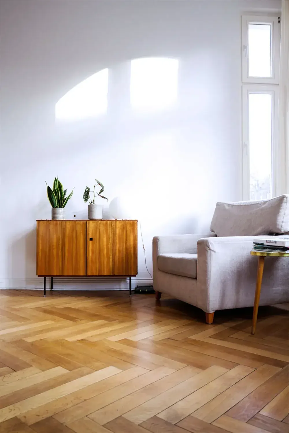 A living room with a wooden cabinet, houseplants, a light-colored sofa, and a window letting in natural light.