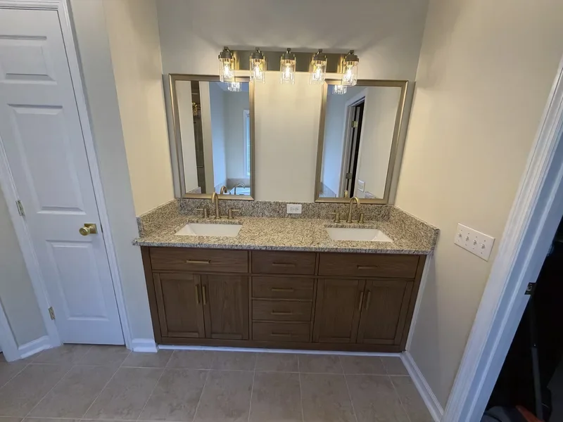 A bathroom vanity with two sinks, granite countertop, wooden cabinet, large mirror, and a row of three light fixtures.