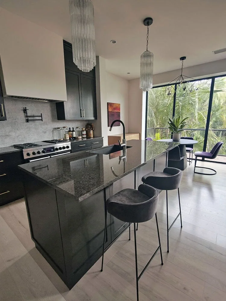 Modern kitchen with black cabinets, a black island with a granite countertop, and barstools. Behind, there are large windows with greenery outside, and hanging pendant lights over the island.