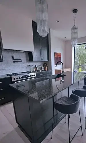 Modern kitchen with black countertops, black cabinets, a stove, and a large window with a view outside. Two hanging chandeliers above the island, with three barstools at the counter.
