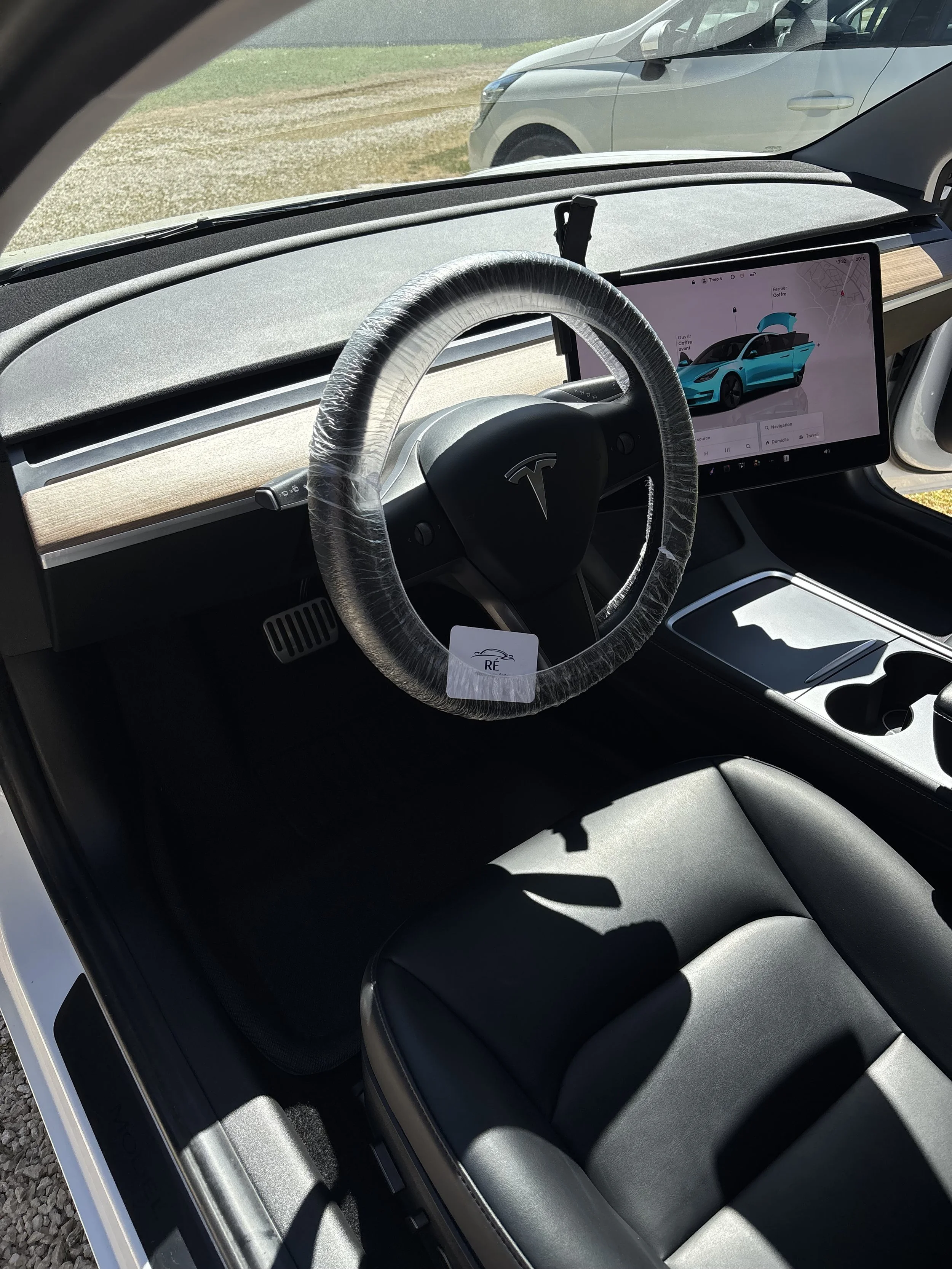 L'intérieur d'une voiture électrique Tesla, vue du siège du conducteur. Un écran tactile montre une voiture électrique bleue avec le capot ouvert, indiquant une opération de chargement ou de maintenance. Le volant est couvert d'un film plastique, et 