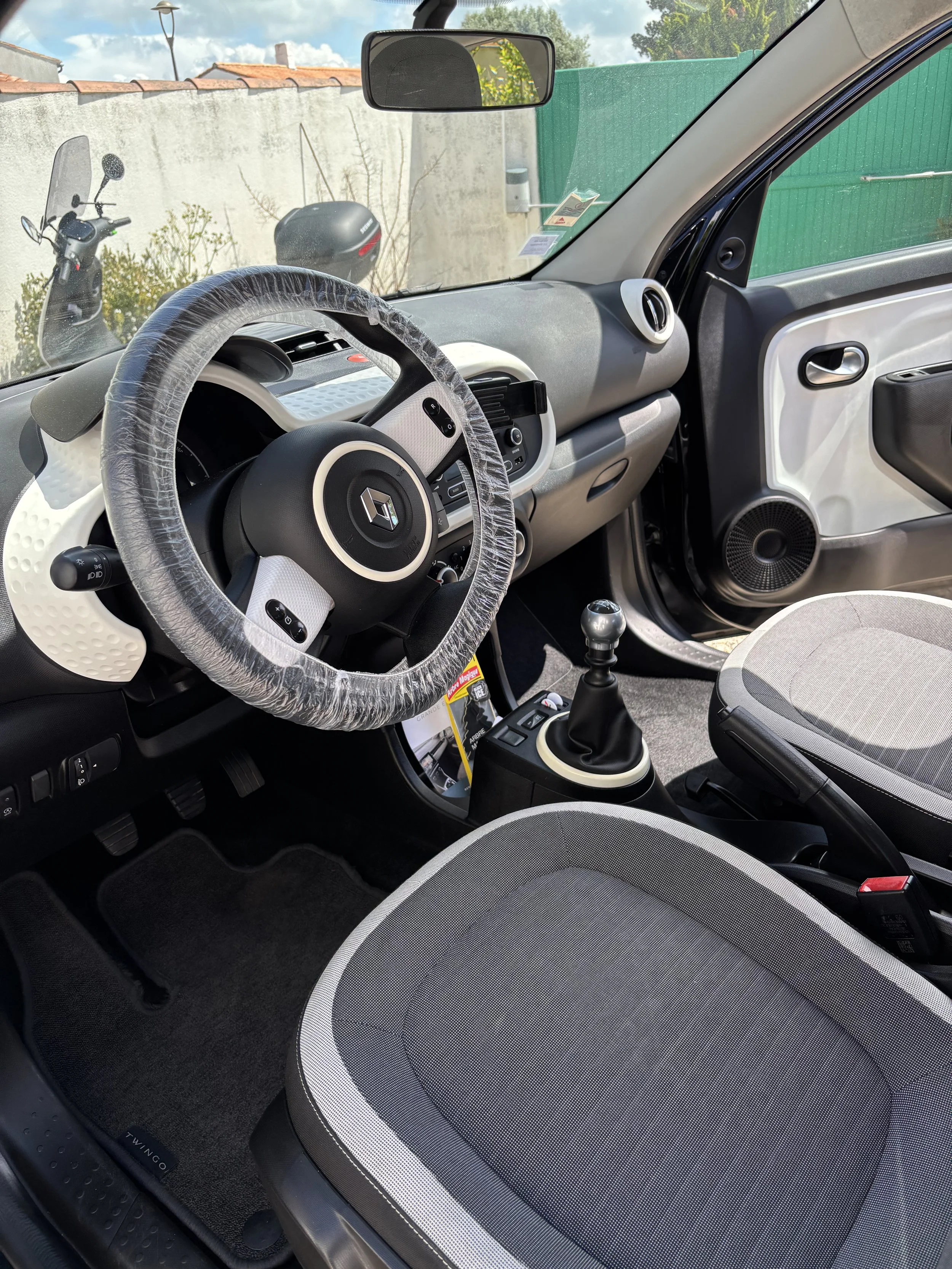 Intérieur d'une voiture Renault moderne, vue du tableau de bord, du volant, des sièges et du levier de vitesses, avec un pare-brise montrant un scooter et un mur extérieur.