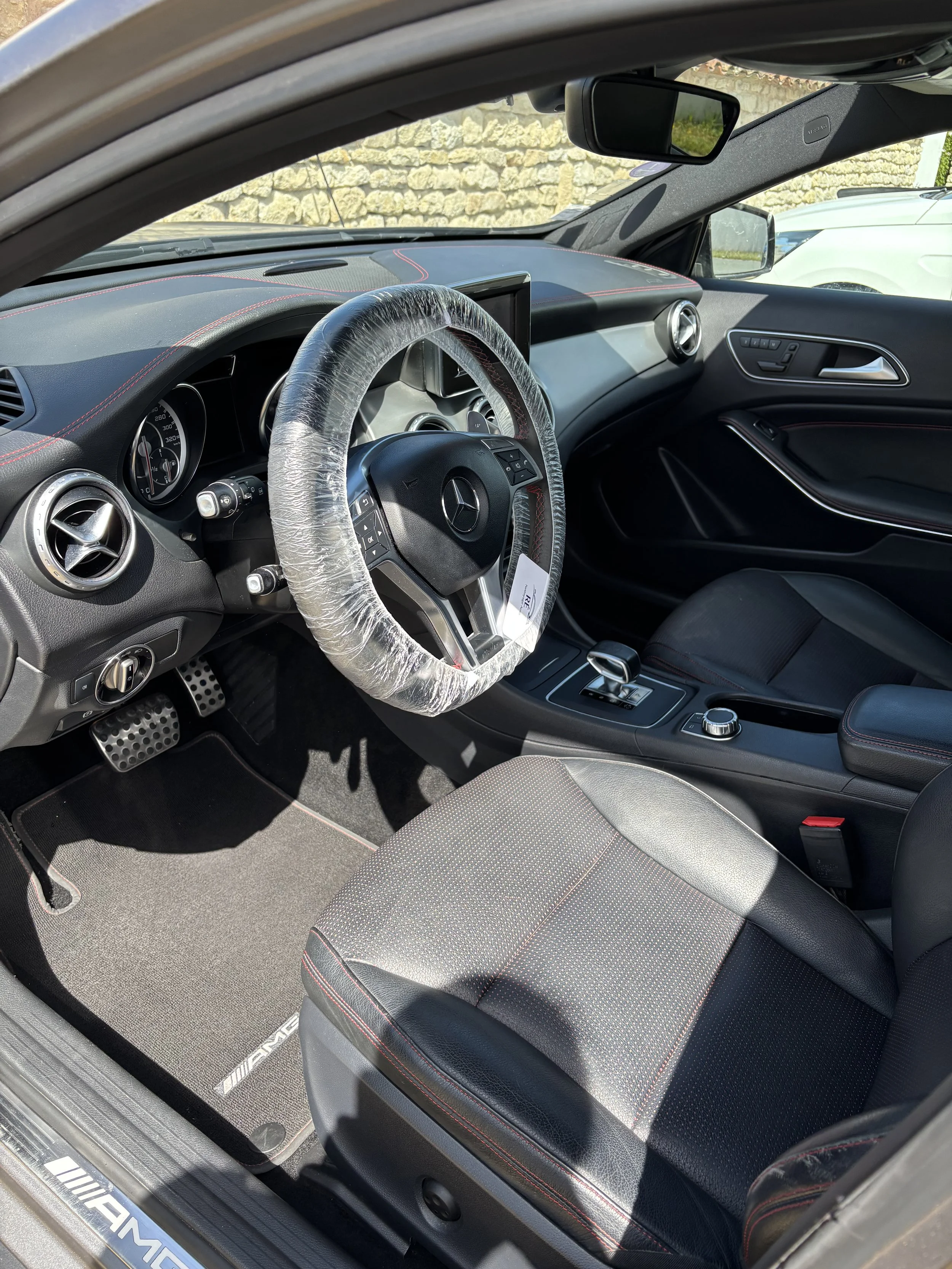 Intérieur d'une voiture Mercedes AMG avec volant, tableau de bord, siège en cuir noir avec coutures rouges, tapis AMG, et volant encore emballé dans un film plastique.