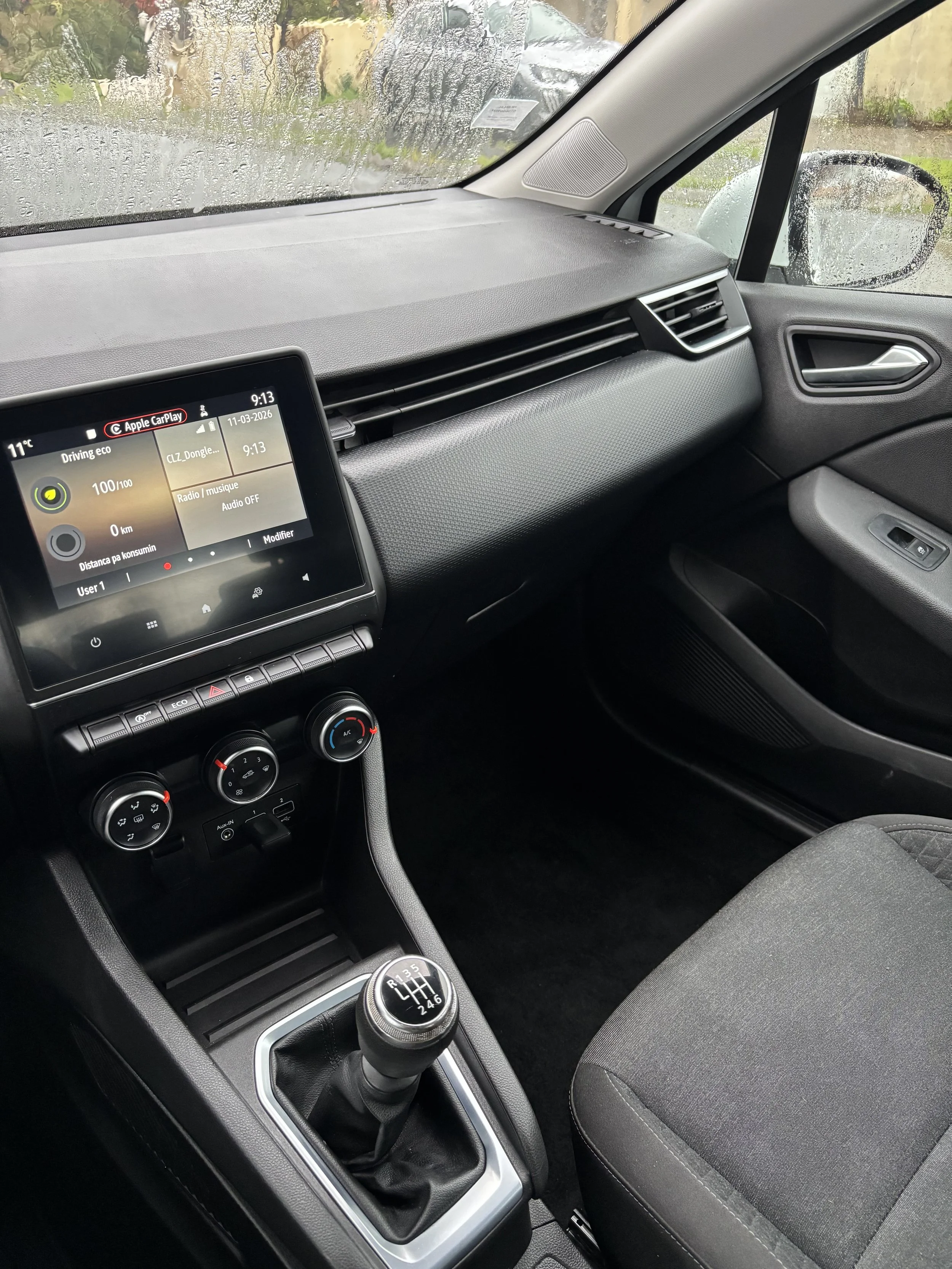 Intérieur d'une voiture, vue du tableau de bord, volant, et levier de vitesse. La fenêtre avant montre une pluie battante dehors.