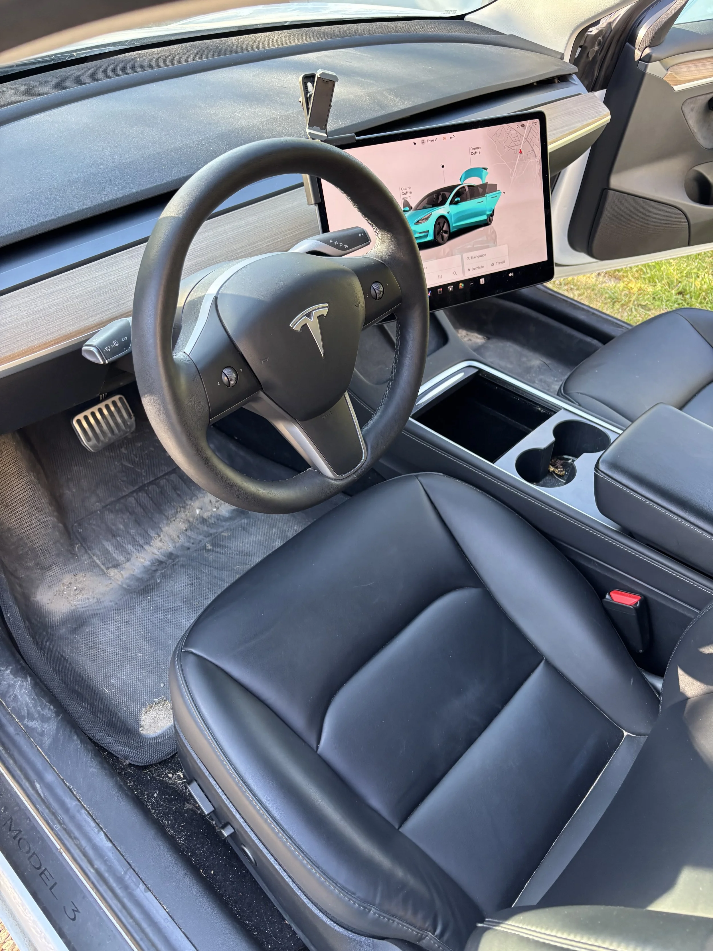 Intérieur d'une voiture Tesla avec un tableau de bord numérique, une tablette tactile affichant la conception d'une voiture électrique bleue, et un couvre-volant, dans un environnement extérieur.