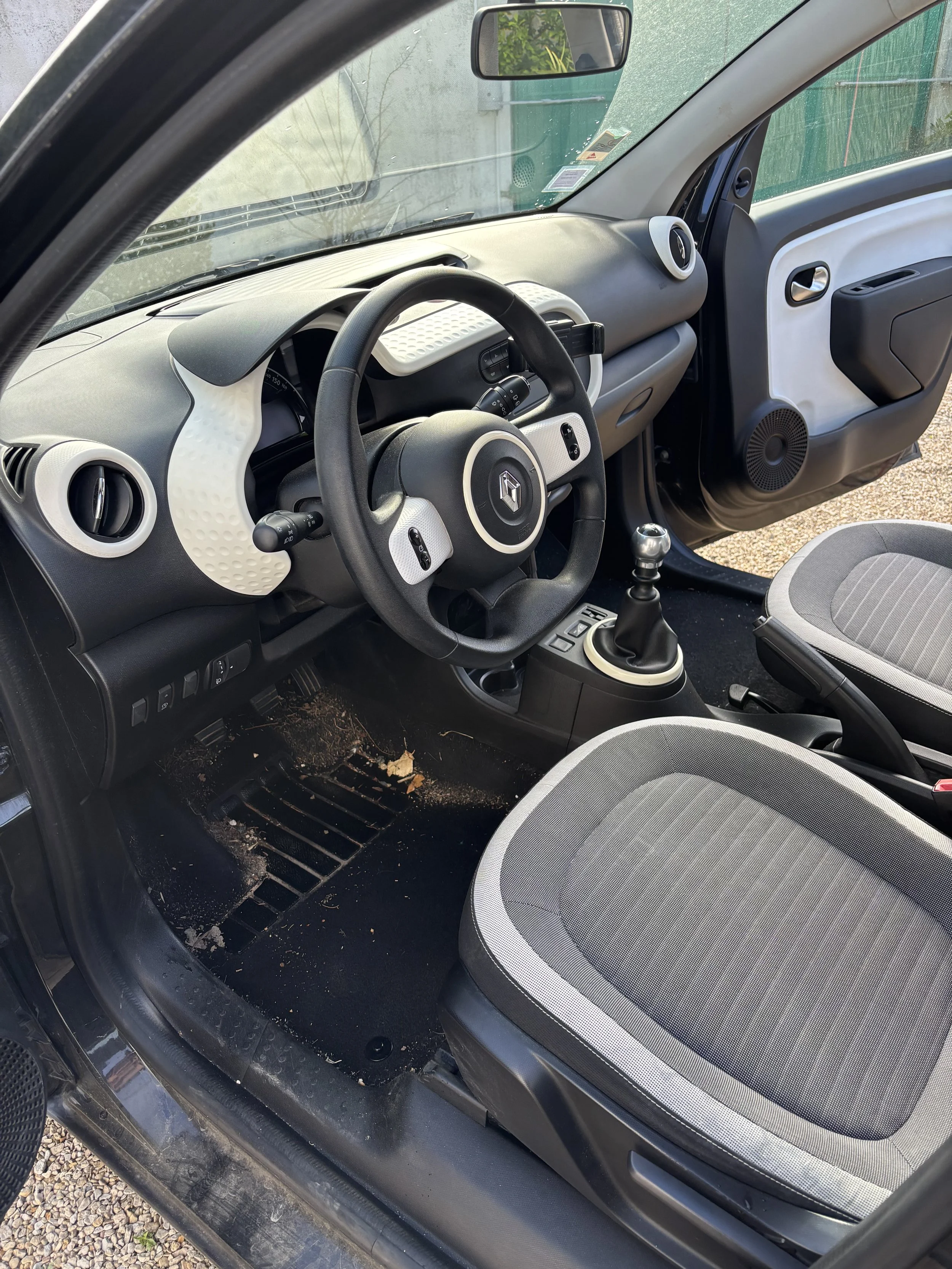 Intérieur d'une voiture compacte avec tableau de bord moderne, volant avec logo Renault, levier de vitesses manuel, et siège en tissu gris à motifs. La porte côté conducteur montre un haut-parleur et un panneau avec un bouton.