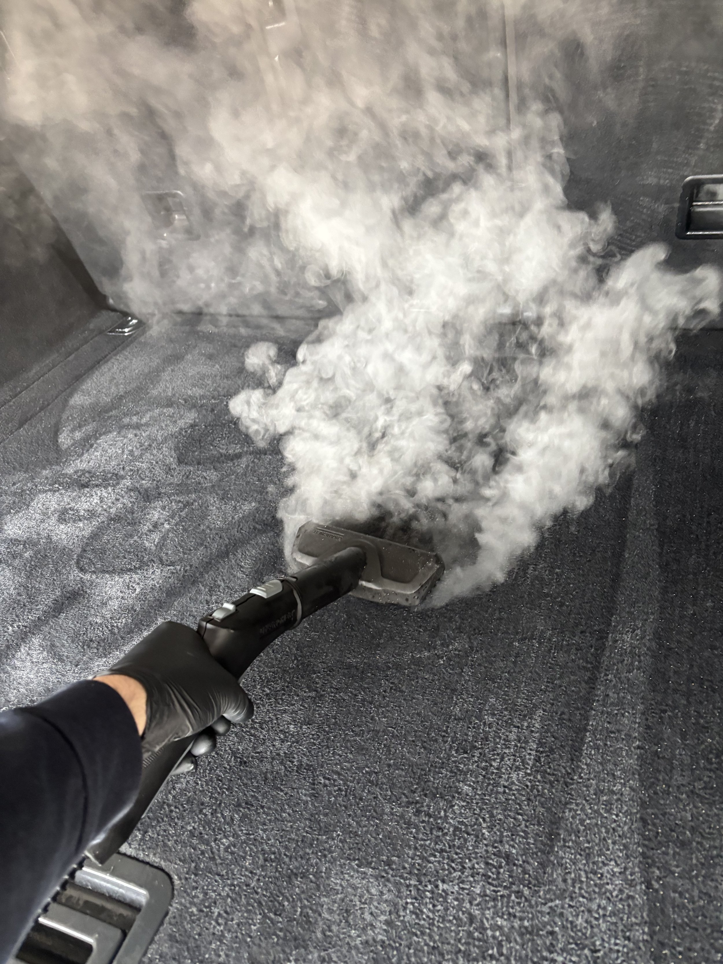 Une personne portant des gants noirs utilise un appareil pour désinfecter ou nettoyer un tapis de voiture, avec de la vapeur ou de la fumée qui s'élève du dispositif.