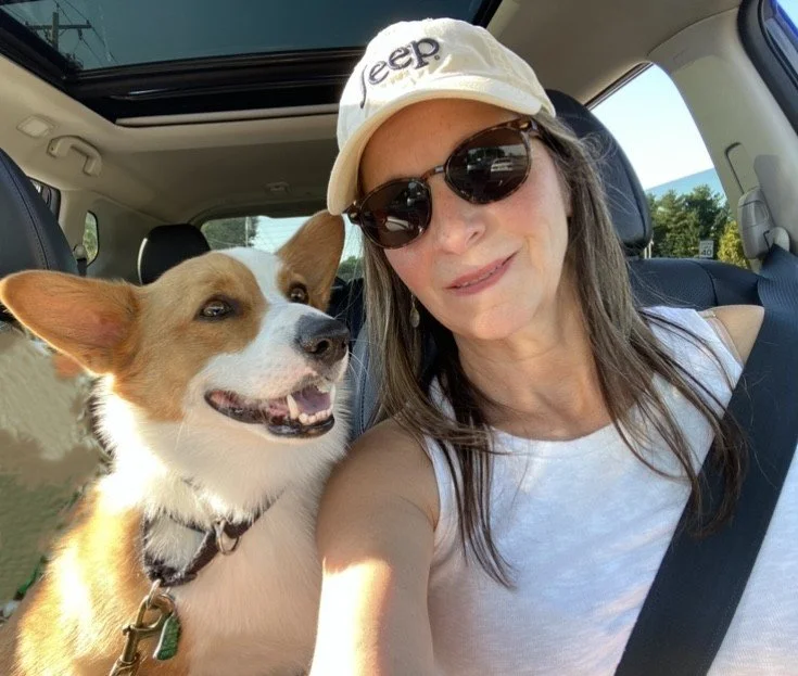 A woman wearing a white Jeep baseball cap, sunglasses, and a white tank top taking a selfie in a car, with a happy brown and white dog sitting next to her.