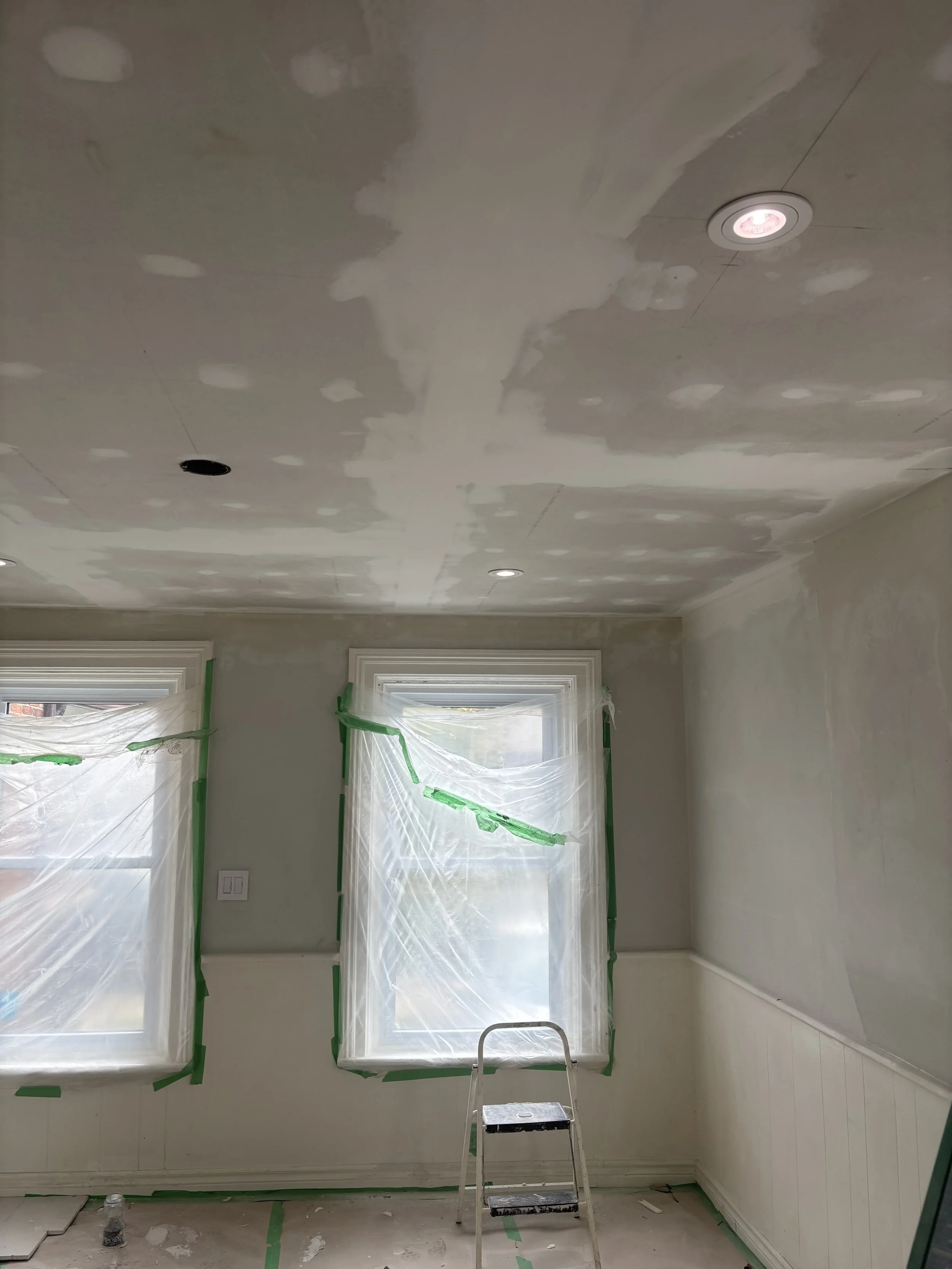 A room with partially painted or textured ceiling and unpainted walls. Two windows covered with plastic and painter's tape. A small step ladder in the center. Light fixtures installed in the ceiling, one with a pink light.