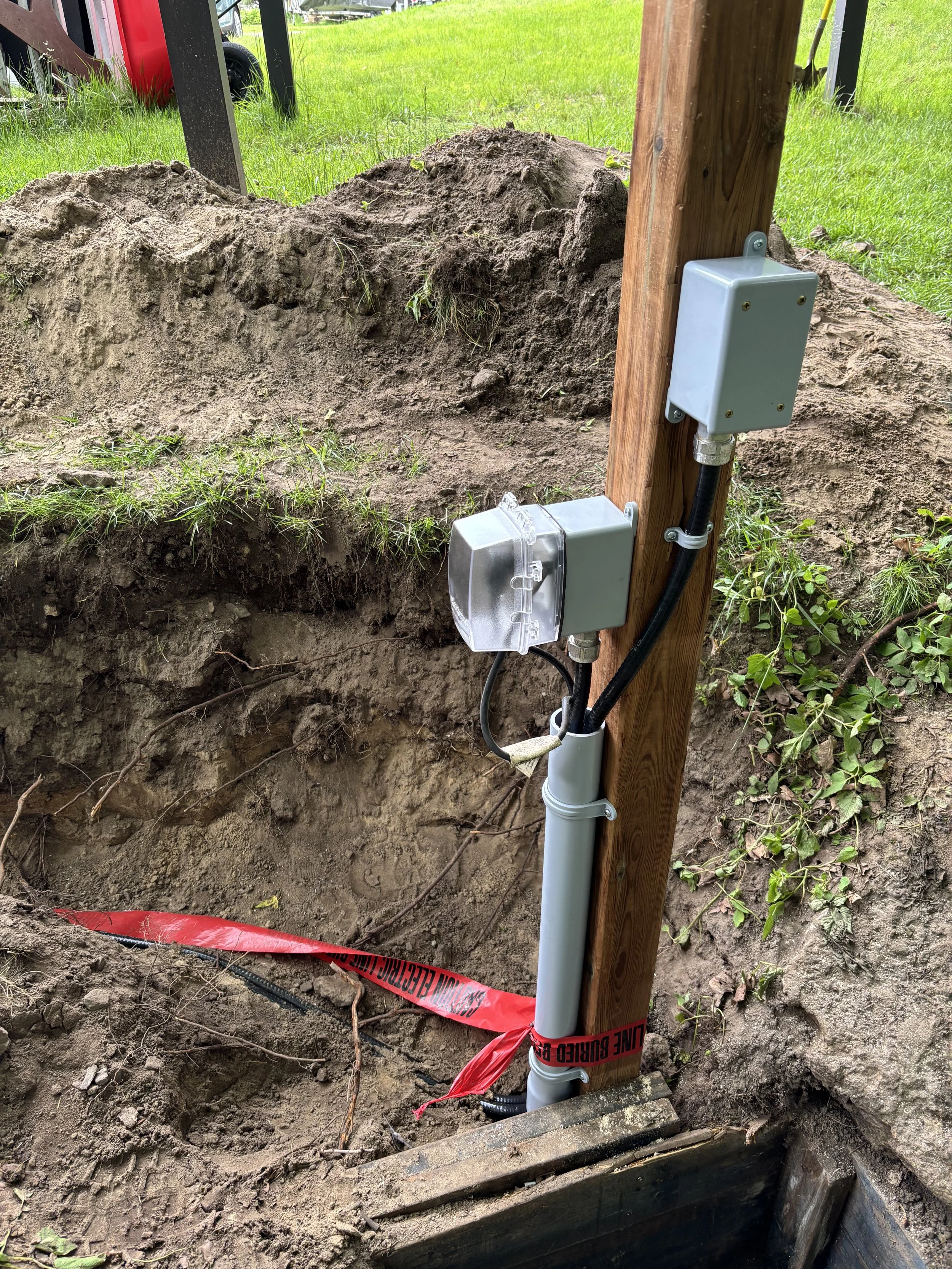 Underground utility electrical box mounted on a vertical wooden post beside a white PVC underground pipe with black electrical wiring, all surrounded by dirt and grass, with red warning tape on the ground.