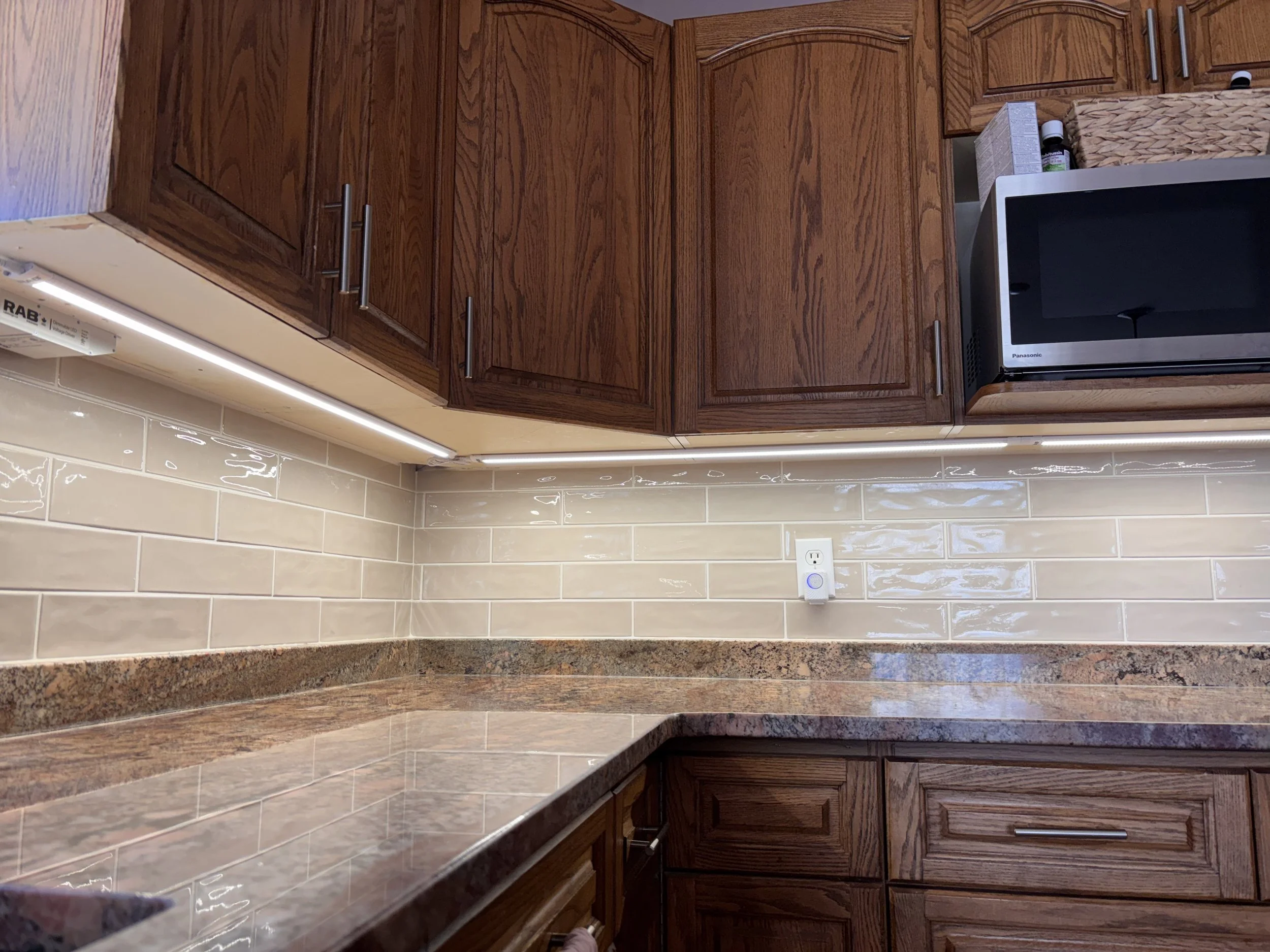 View of kitchen cabinets with a microwave on the right, beige tiled backsplash, stone countertop, and a white electrical outlet.