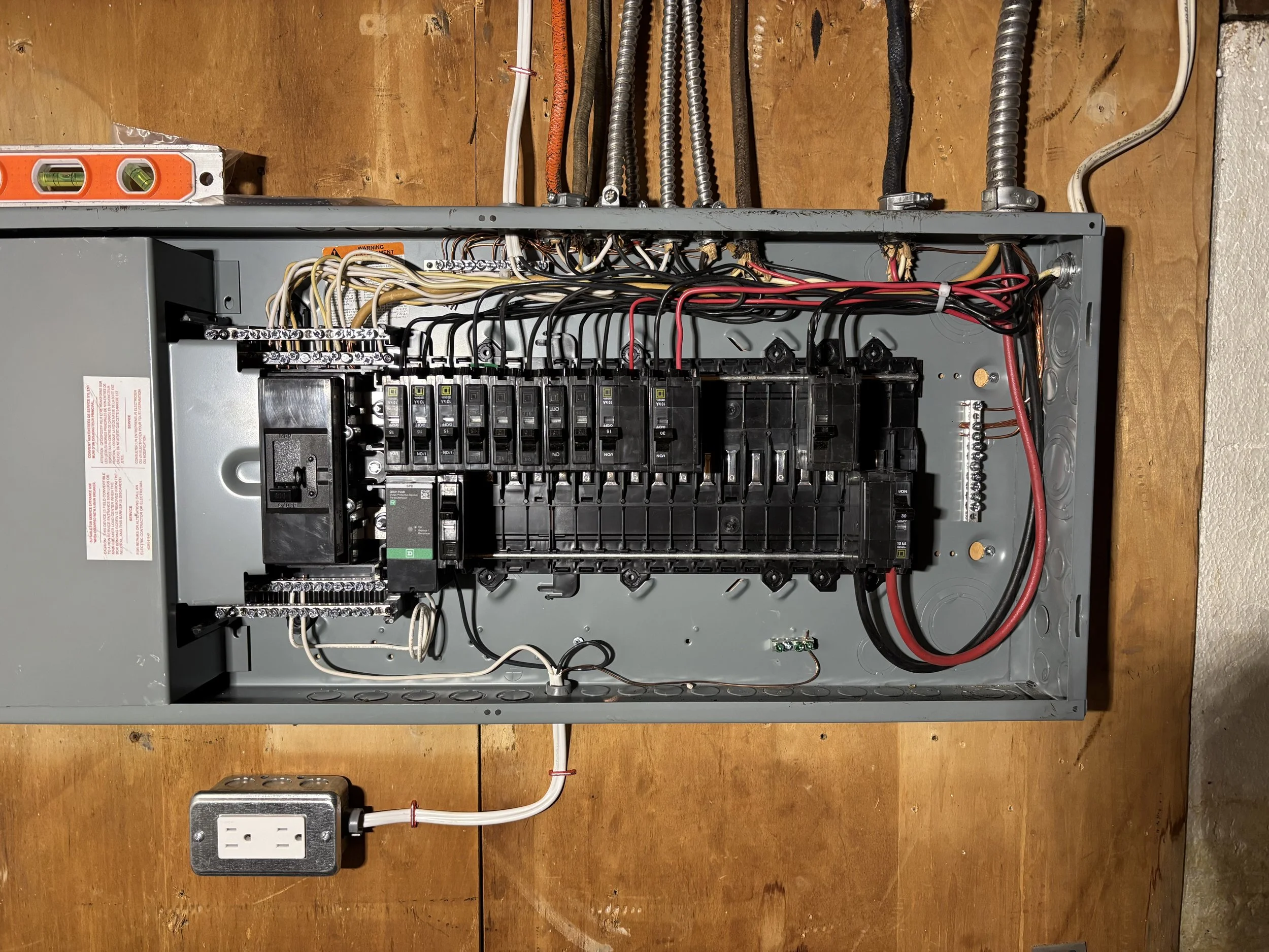 Open electrical panel with various wires and circuit breakers mounted inside on a wooden surface.
