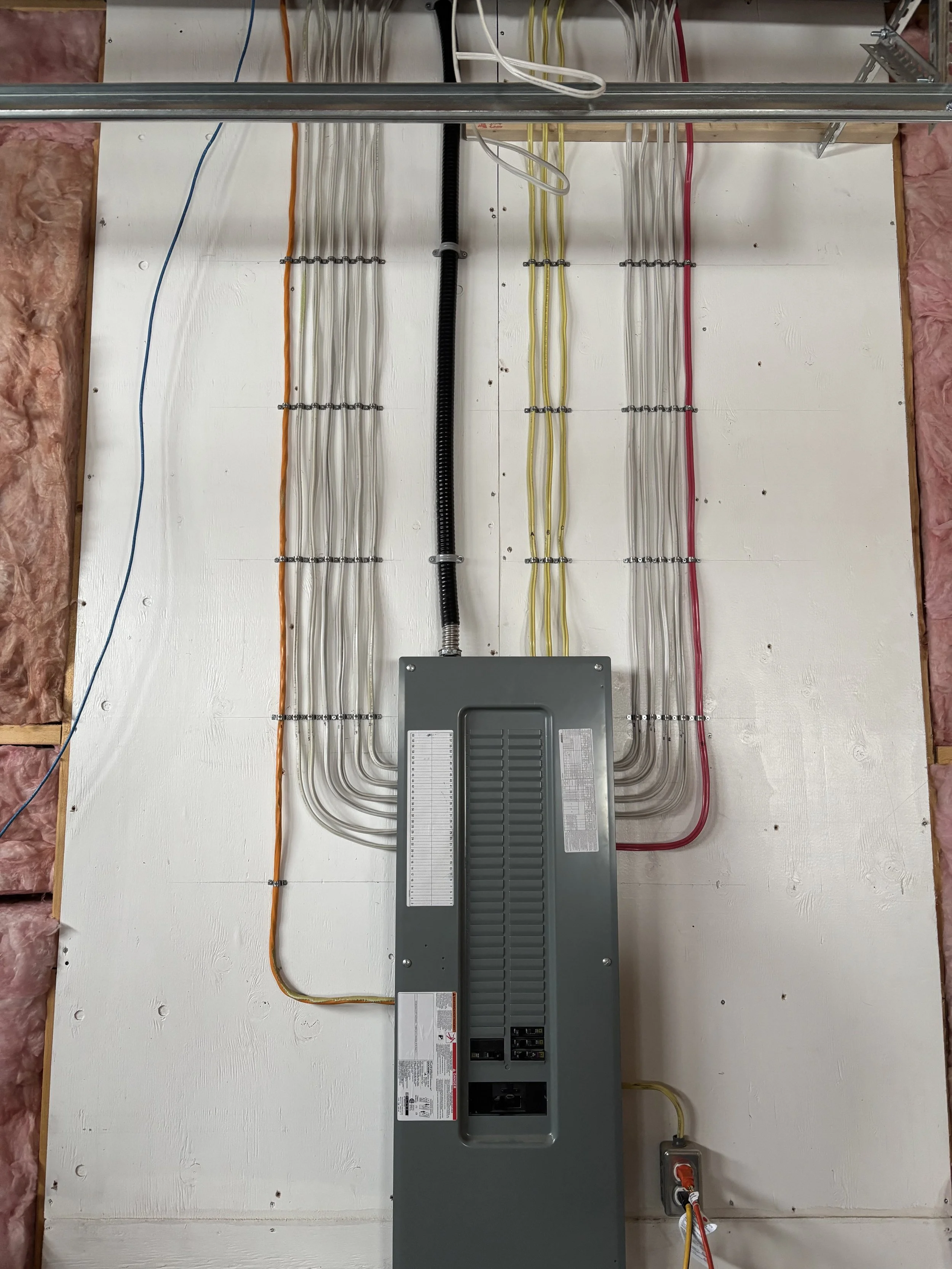 Electrical panel with multiple colored wires attached to a white panel wall, with insulation and construction materials visible on the sides.