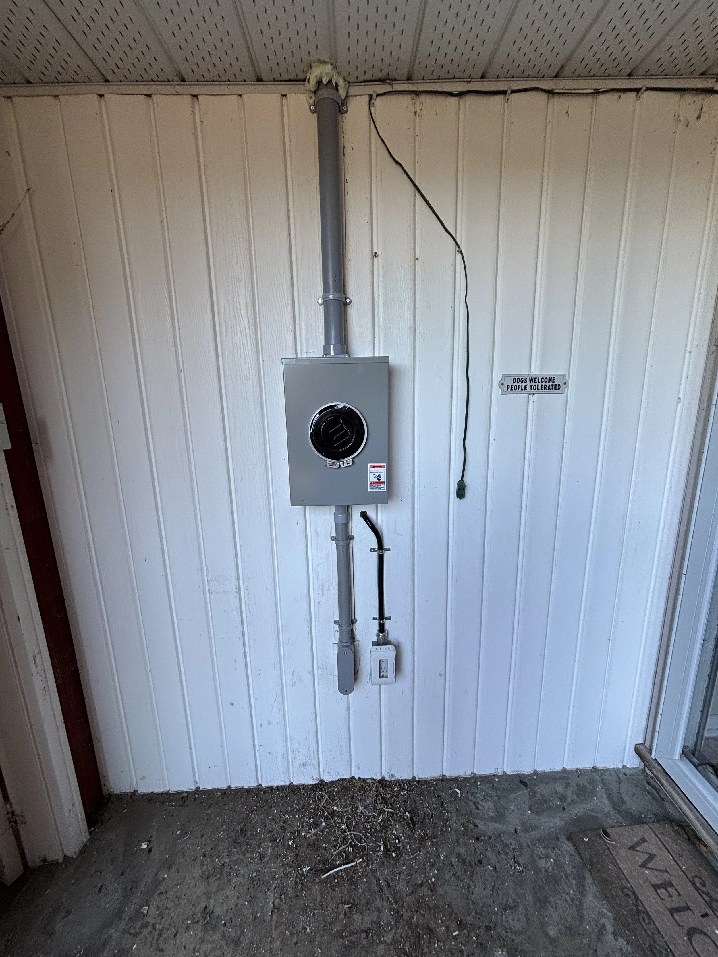The image shows an outdoor wall with a white vertical paneling surface. Mounted on the wall is an electrical box with a circular opening and an attached pipe. A black wire runs from the box to the right side of the wall. There is a small sign that reads 'Dogs Welcome People Tolerated'. The ground is concrete with dirt and debris, and part of a doormat with the word 'WELCOME' is visible on the bottom right.