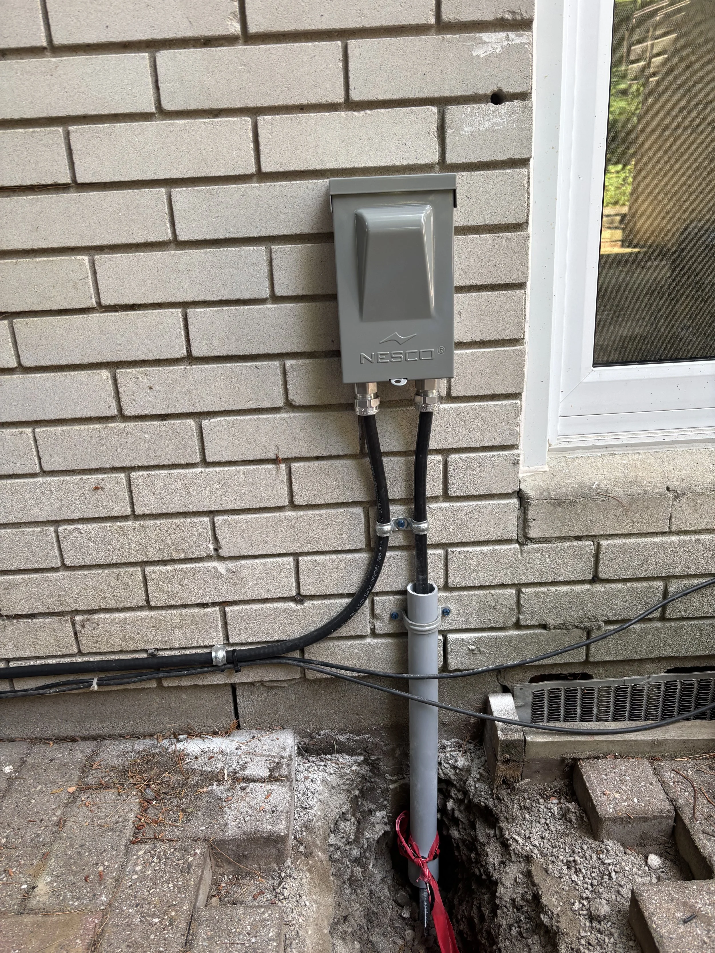 Electrical wiring and conduit pipes installed outside a brick wall, with an electrical box labeled 'NESCO' and part of the wall showing a hole and a window frame.