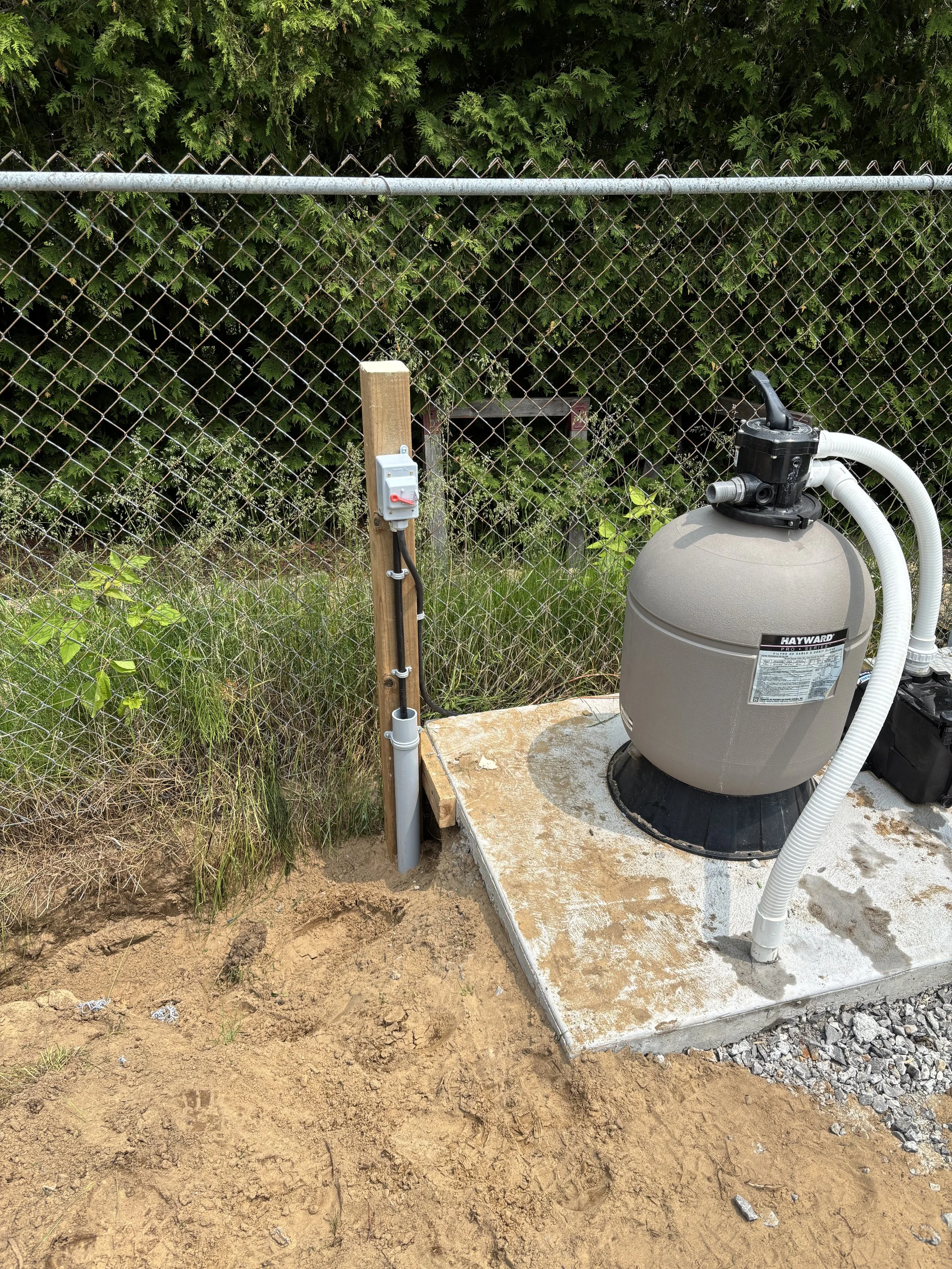 A pool filter system installed outdoors on a concrete slab, next to a chain-link fence and green shrubbery.