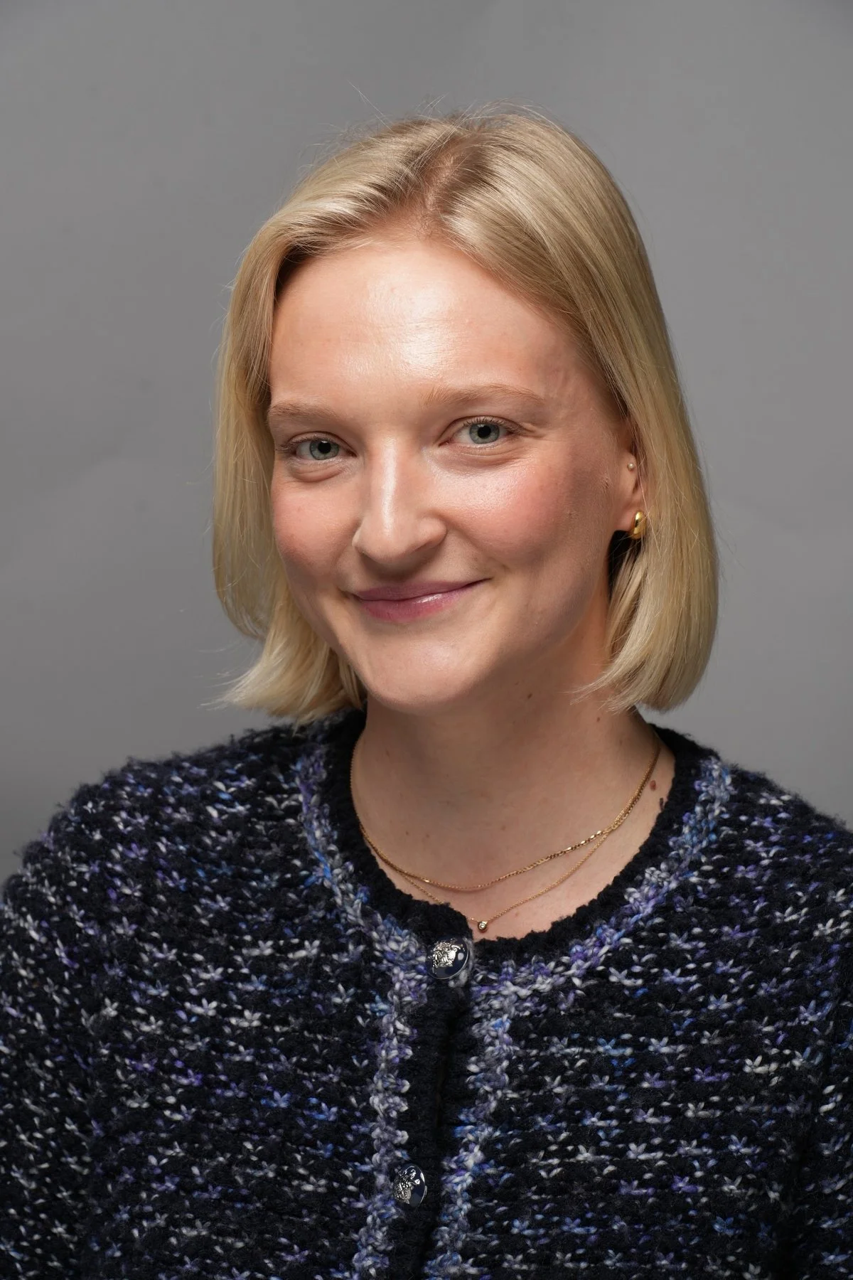 Close-up portrait of a smiling woman with blonde hair, wearing a dark textured sweater with buttons and gold jewelry against a neutral grey background.