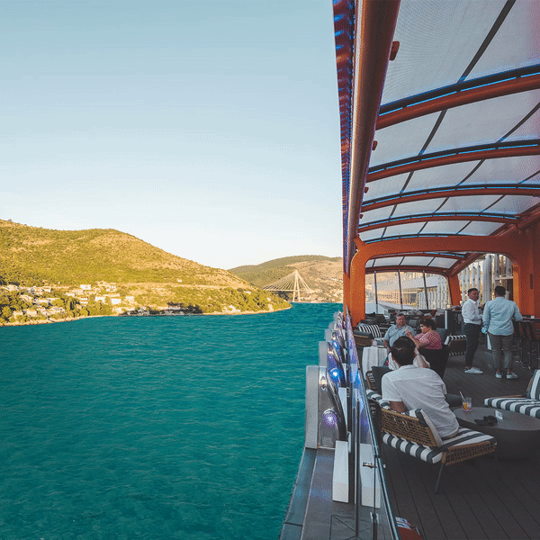 People relaxing on the deck of a cruise ship with a view of the water and landscape with hills and a bridge in the background.