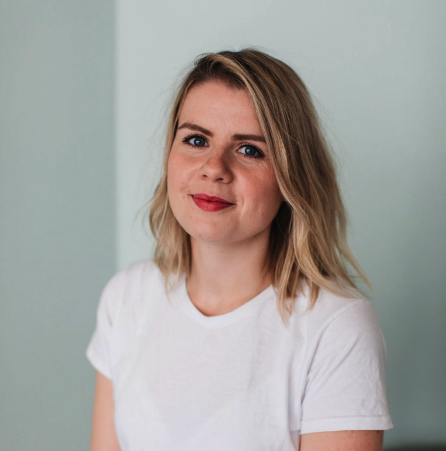 A young woman with shoulder-length blonde hair, blue eyes, and light makeup, smiling slightly, wearing a white T-shirt, standing in front of a light-colored wall.