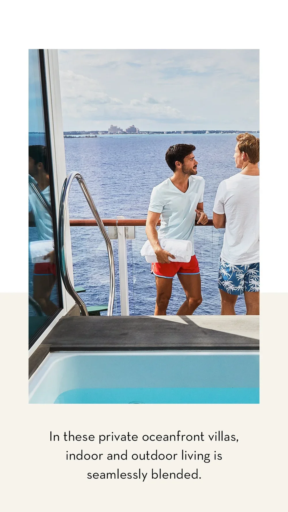 Two men in swimwear having a conversation on the balcony of a private oceanfront villa, with a view of the water and distant buildings in the background.