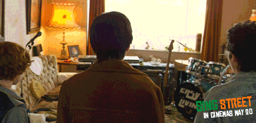 Three people sitting in a music studio with a drum set, microphone, and mixing equipment, facing a window with sunlight coming through.