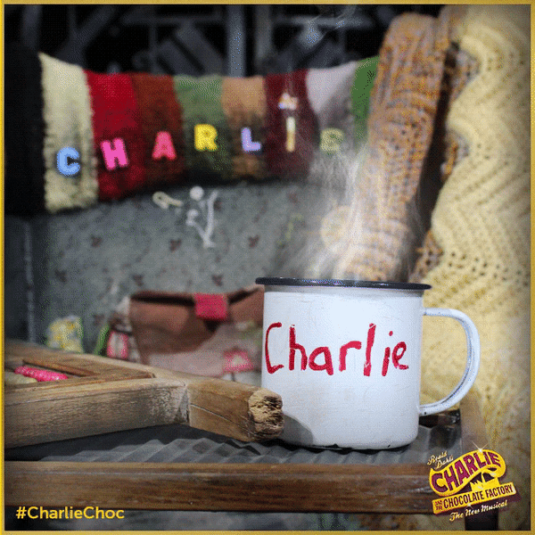 A steaming mug with the name 'Charlie' written on it, placed on a wooden tray. In the background, colorful knitted letters spell out 'CHARLIE' on a textured, multicolored blanket. The scene appears to be cozy and outdoors.