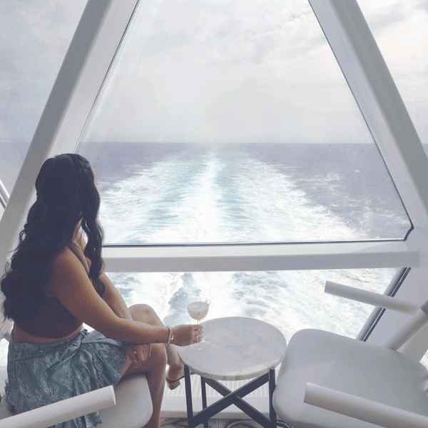 A woman with long dark hair sitting inside a modern vessel's interior, looking out at the ocean through a large window, holding a glass of wine, with the wake of the boat visible behind her.