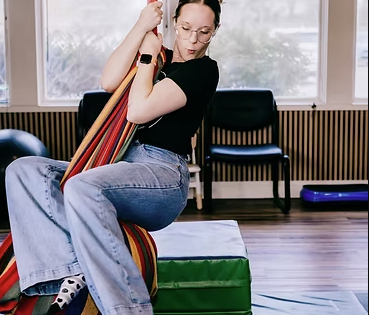  woman sitting on a padded blue gym block holding colorful exercise mats indoors.