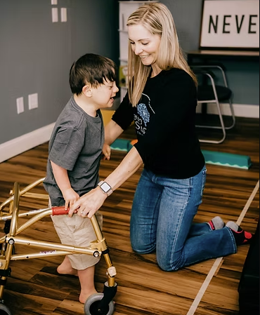 A woman kneeling on the floor helping a young boy with walking using a walker in a room with wooden flooring and a sign that says 'NEVER' in the background.