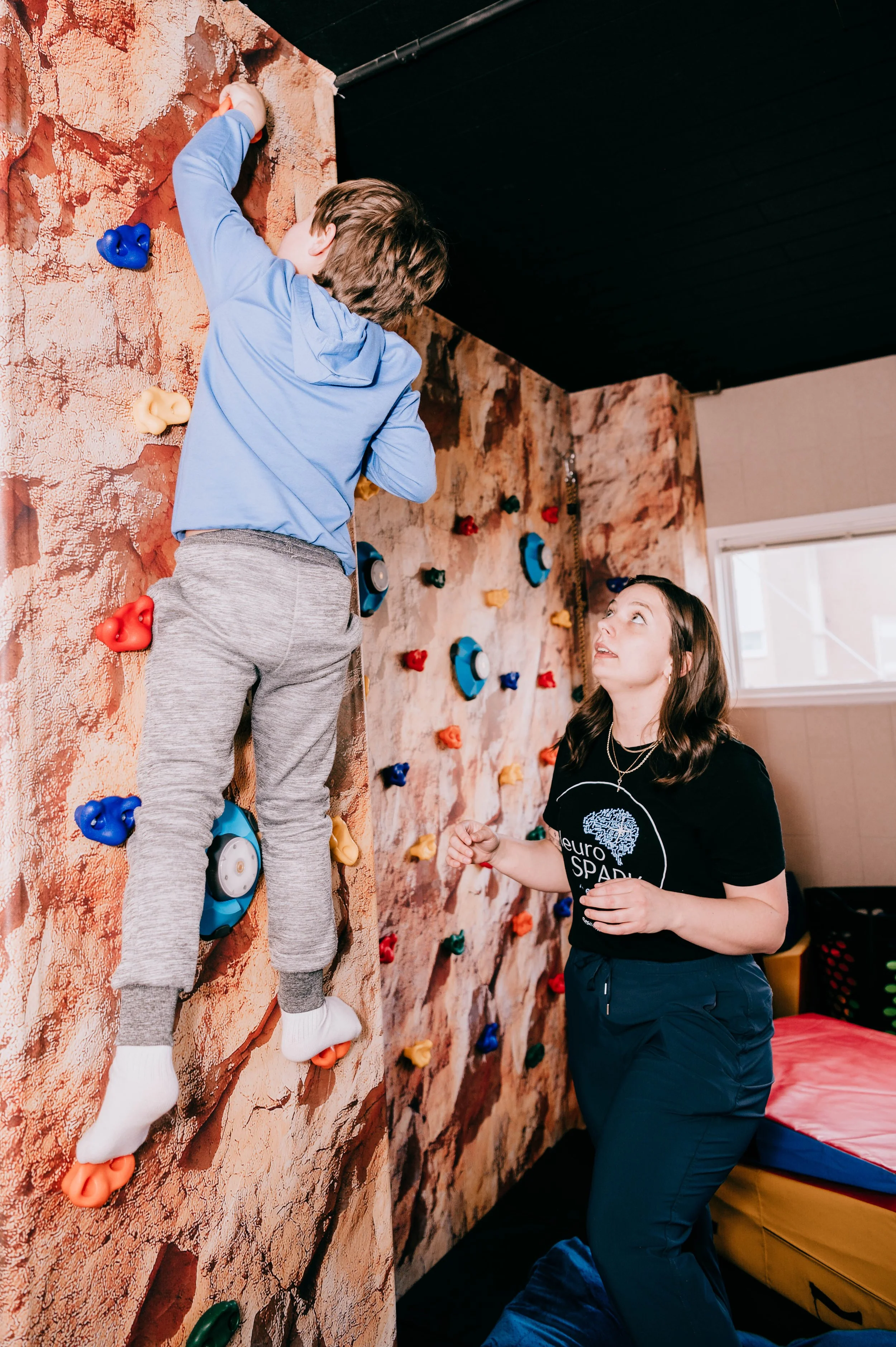 A young boy in a blue hoodie and gray sweatpants climbing an indoor rock wall with colorful holds. A woman watches and encourages him, kneeling near the base.