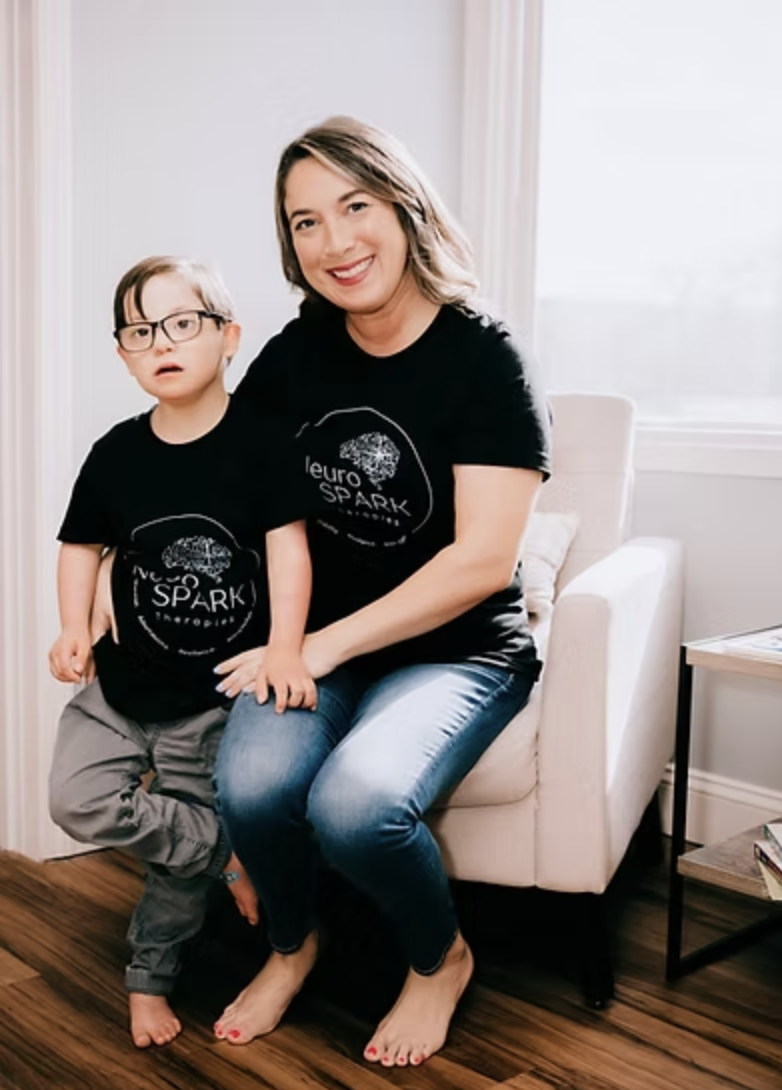 A smiling woman and a young boy sitting together on a white armchair in a bright room. Both are wearing matching black T-shirts with a logo that says "leuro SPARK" and a tree graphic. The woman is barefoot, and the boy is barefoot and wearing glasses.