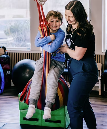 A young boy sitting on a colorful gymnastics block holding onto red and yellow straps, smiling with a woman standing beside him, both smiling, in a room with large windows and gym equipment.
