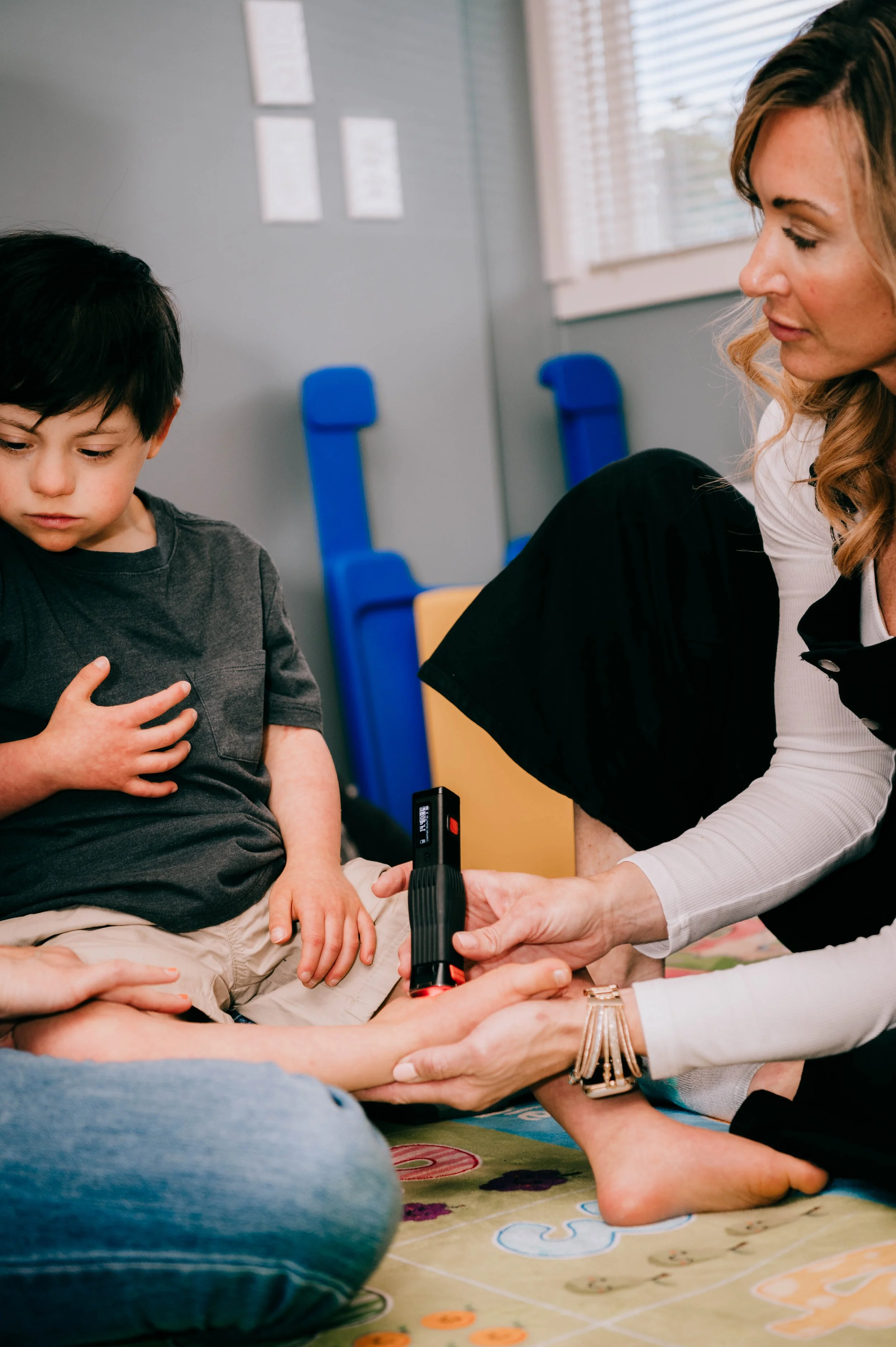 A young boy sitting on a colorful playmat with medical professionals around him, as a woman prepares to use a blood glucose meter on his finger in a well-lit room.