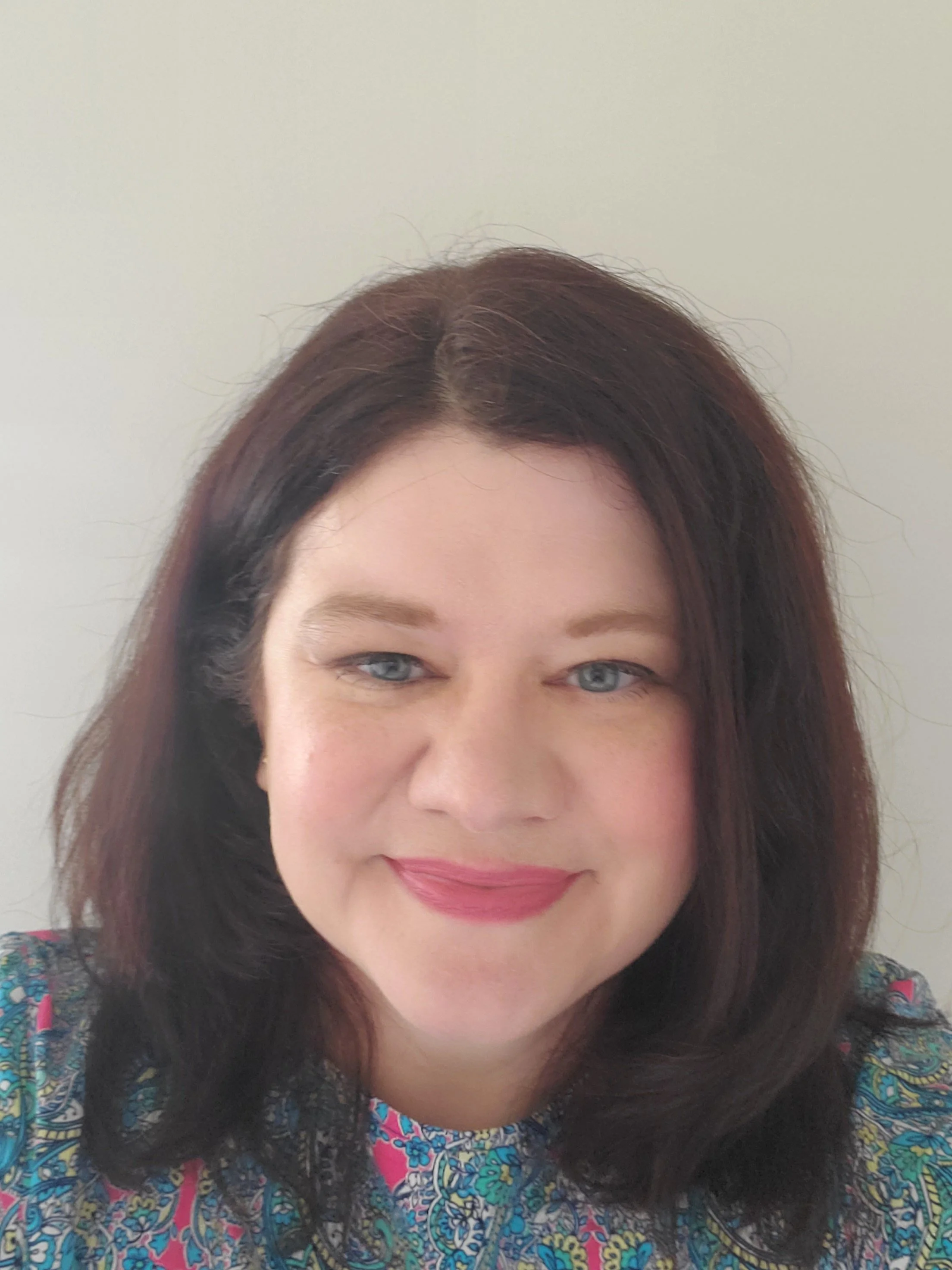 Close-up of a woman with dark brown hair, blue eyes, light skin, wearing a colorful patterned top, smiling, against a plain gray background.
