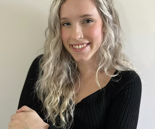 A young woman with long, curly blonde hair, wearing a black ribbed top and smiling at the camera, against a plain background.
